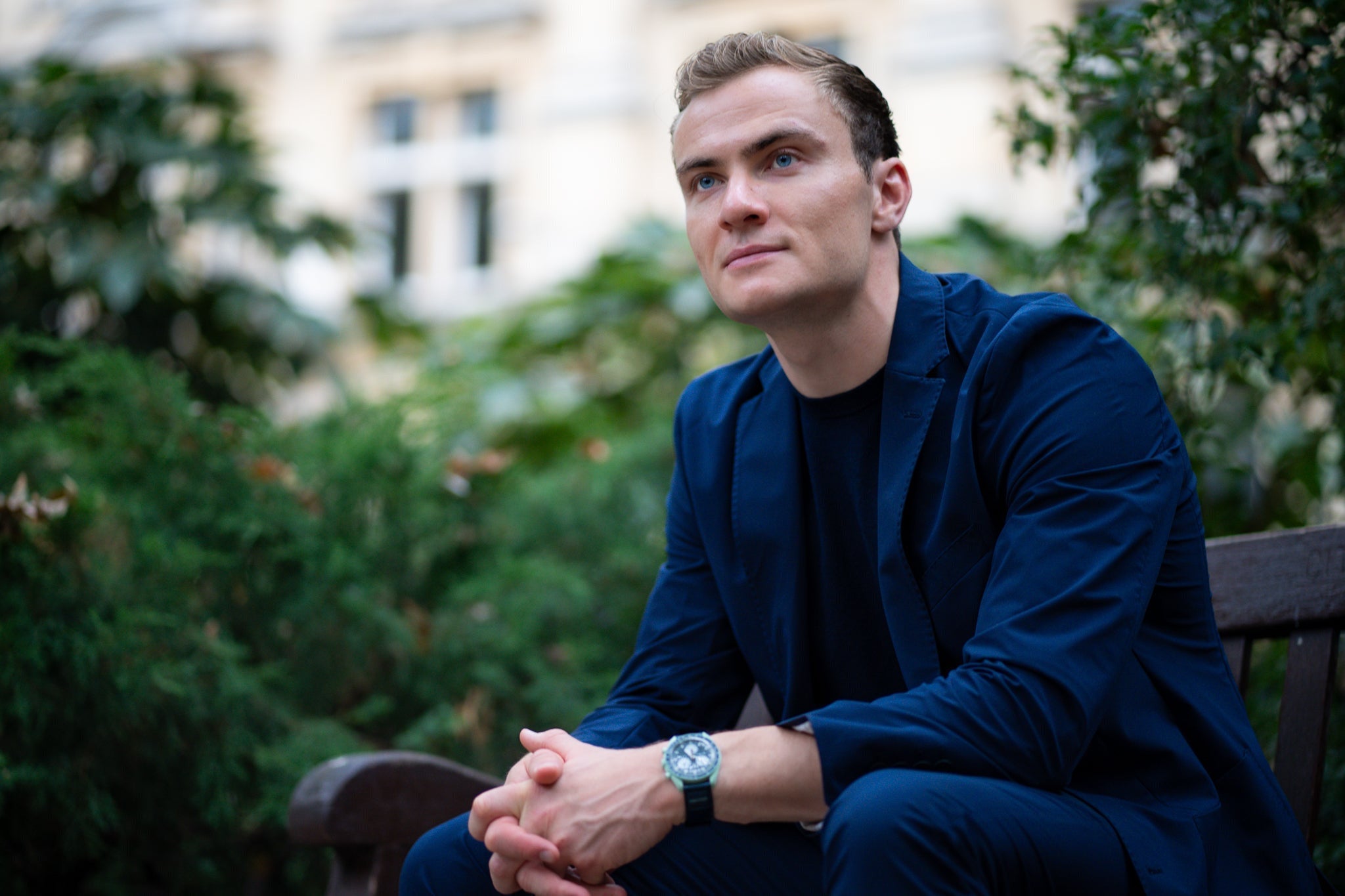 Man in a blue suit sitting on a bench with greenery in the background