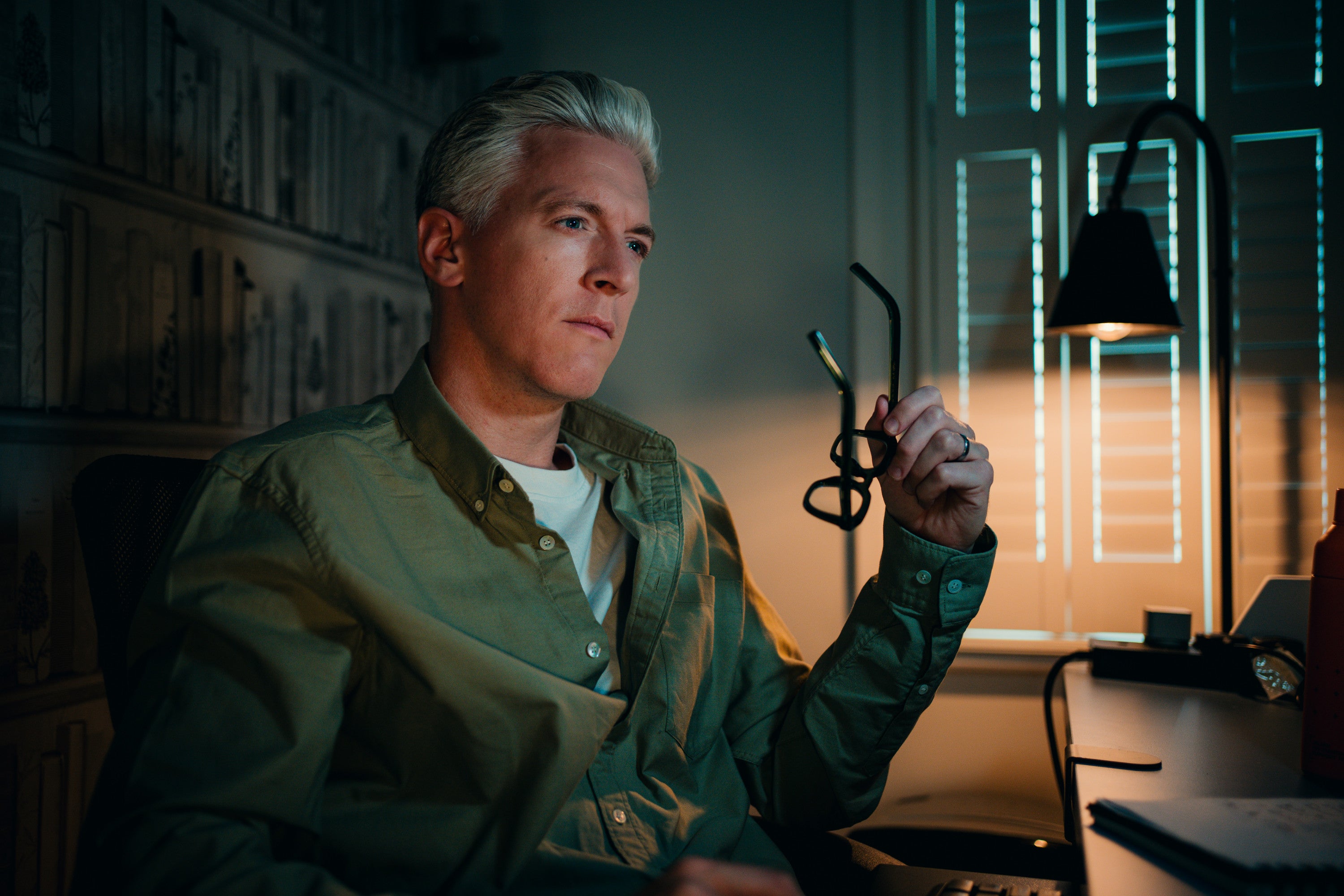 Man holding eyeglasses in a dimly lit room with a window in the background