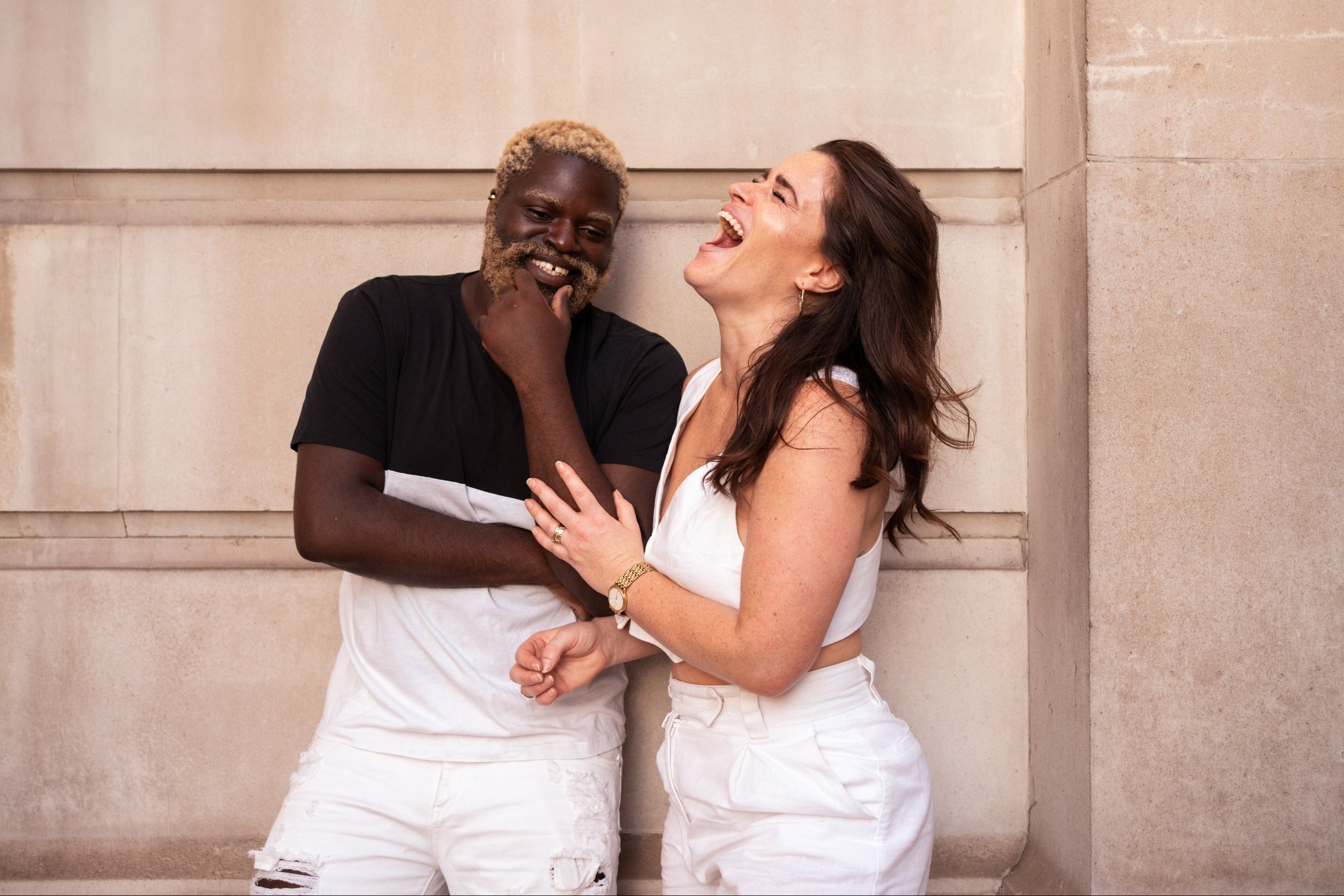 Two people laughing against a stone wall.