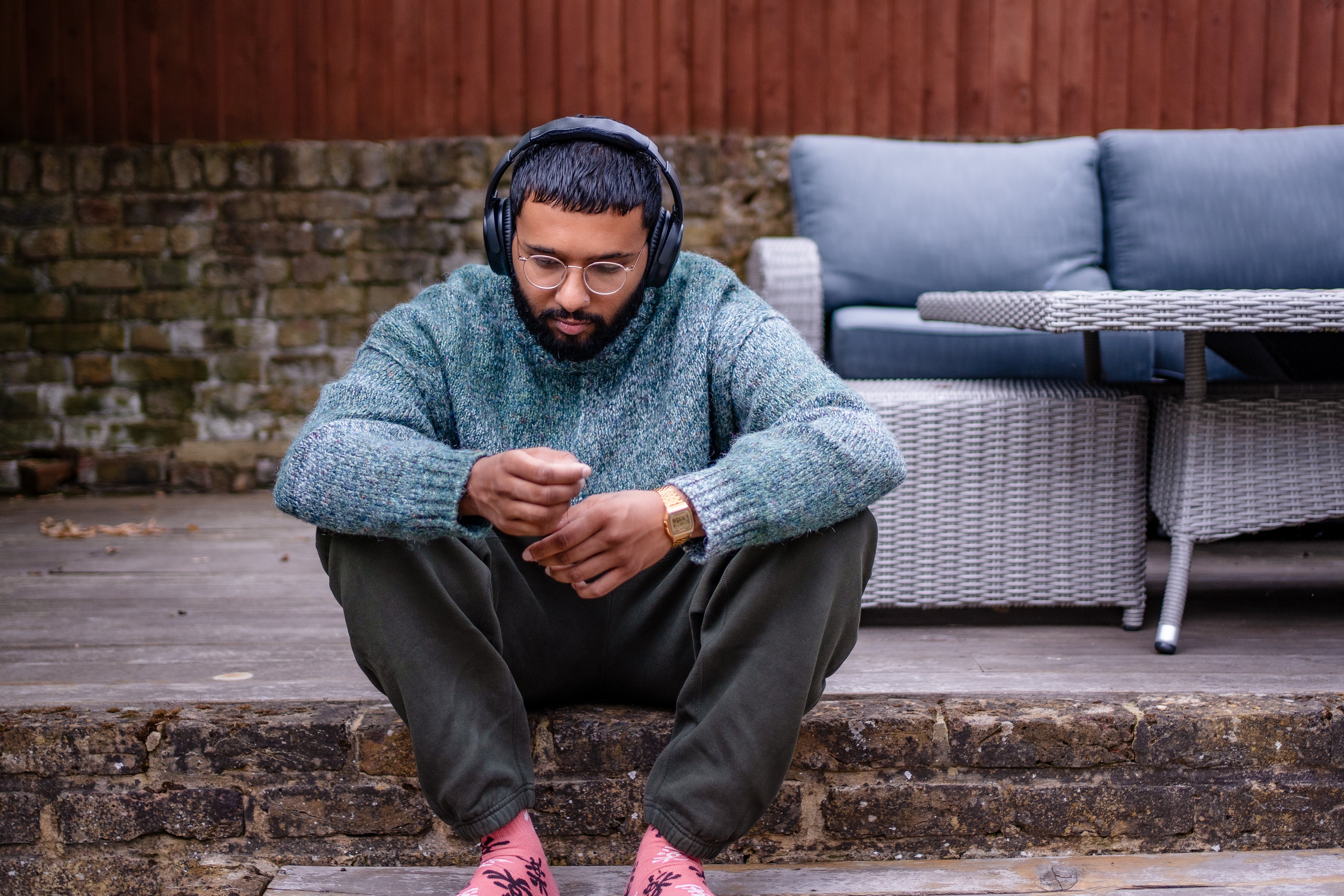 Man sitting on steps wearing headphones and a green sweater, with outdoor furniture in the background.