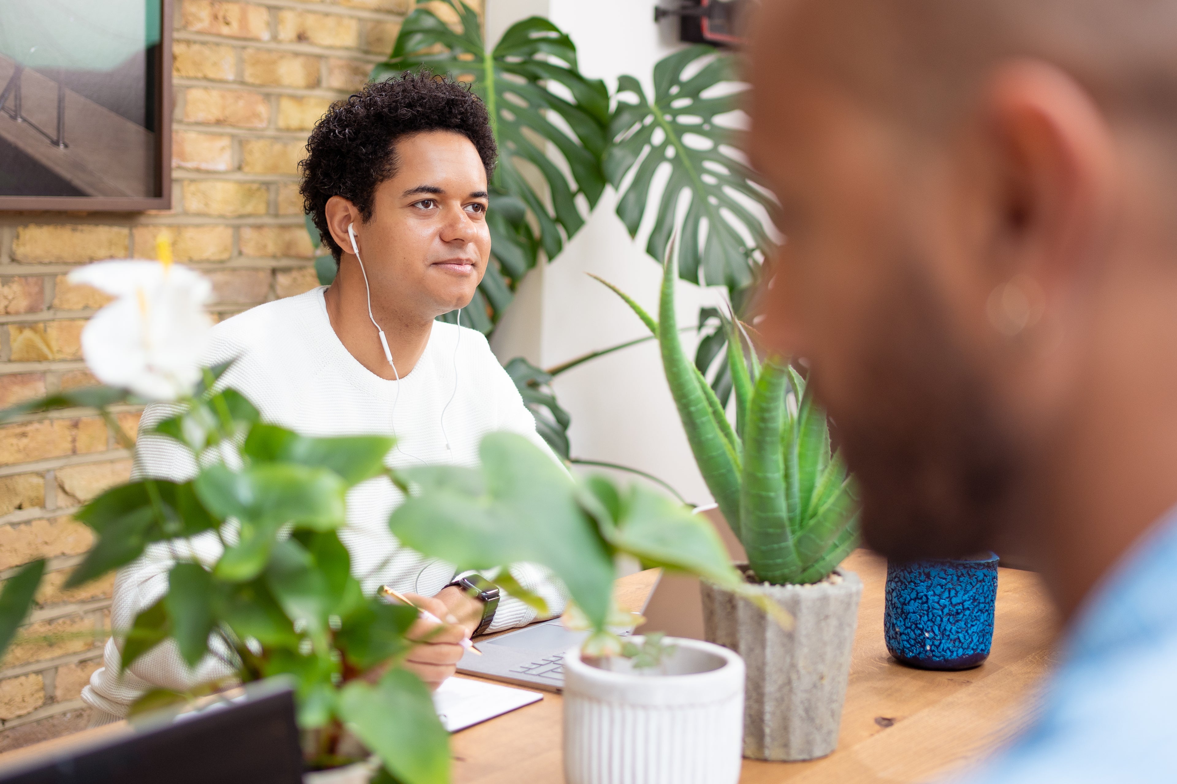 Man sitting at a desk with plants and another person partially visible