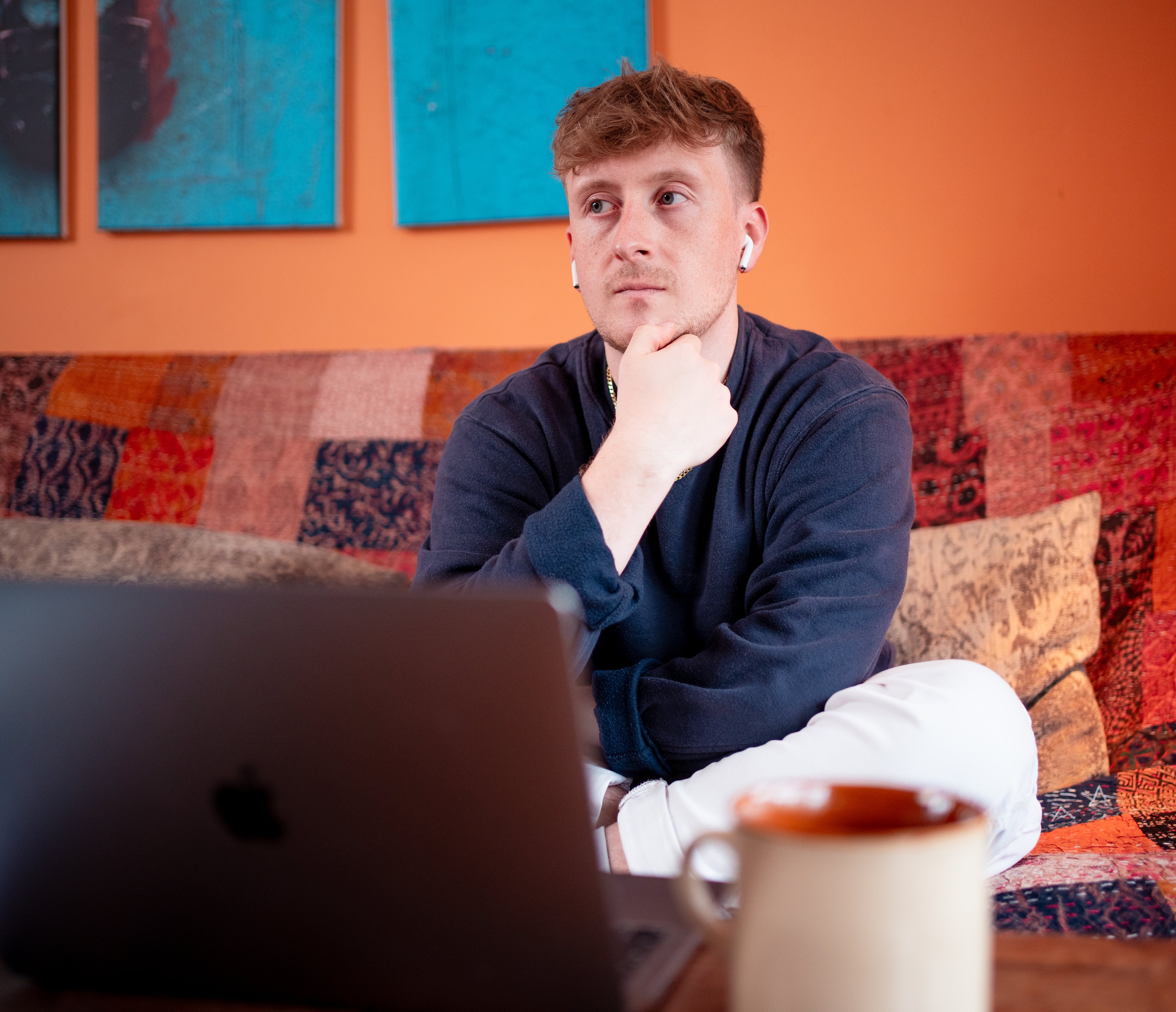 Man sitting on a colorful couch with a laptop and coffee mug, looking thoughtful.